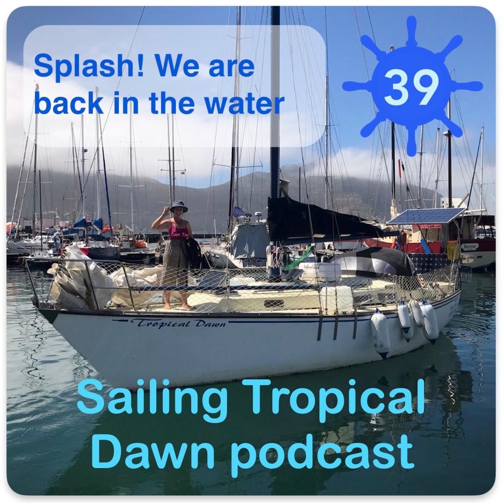 Image of a sailboat with a woman standing on the deck (Kay) text at bottom: Sailing Tropical Dawn podcast; top left: Splash! We are back in the water and top right a sailboat steering wheel with the number 39 inside